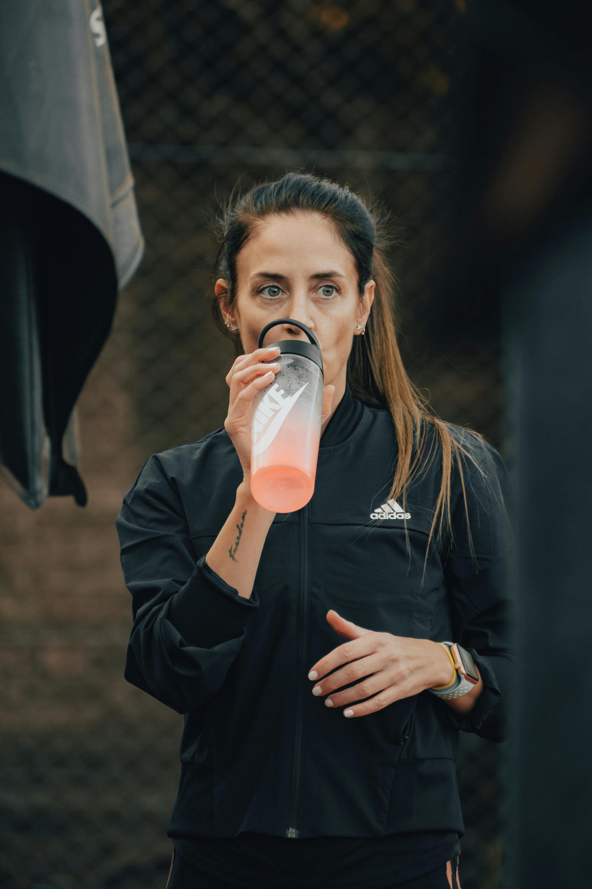 Athletic woman in black sports jacket hydrating with a bottle outdoors.