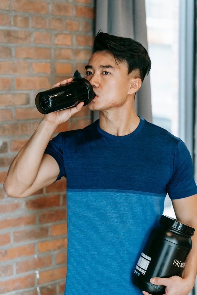 Confident young Asian male athlete with dark hair in activewear drinking water while standing near brick wall in gym with protein powder container