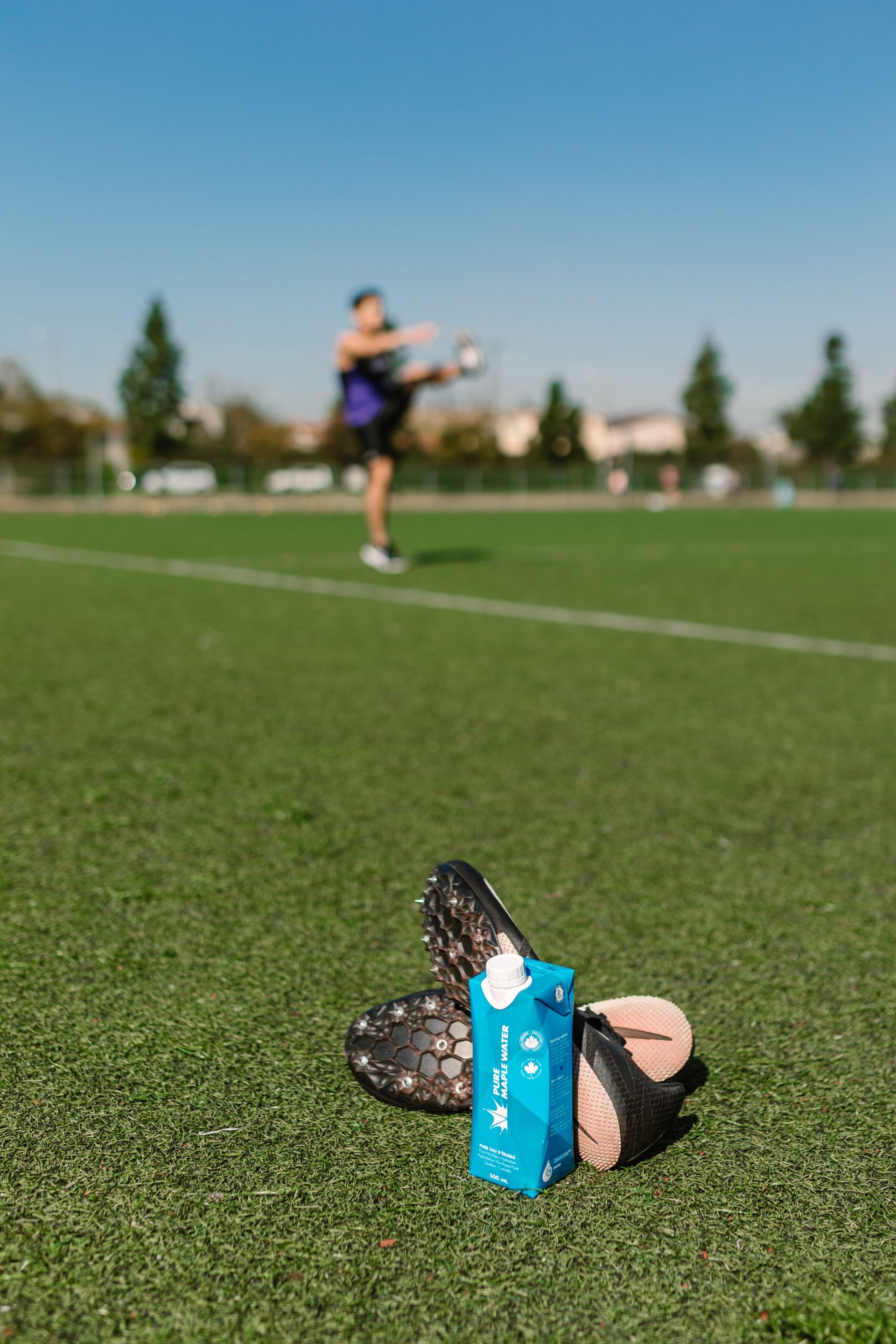 Close-up of soccer cleats and an energy drink with player in background on sports field.